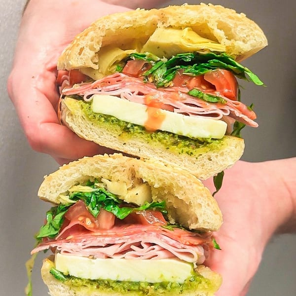 Hands holding a large Italian sandwich, cut in half, filled with meats, cheese, and vegetables.