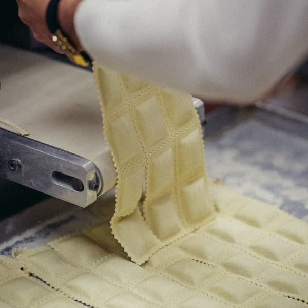 Hands carefully separate freshly made ravioli from a pasta machine, showing rows of square pasta.