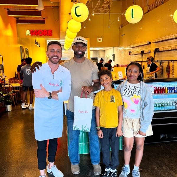 A chef, a man, and two children pose with a Pasta Via bag in a brightly lit restaurant.