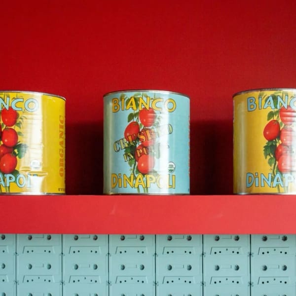 Three colorful Bianco DiNapoli tomato cans on a red shelf above a wall of light blue cassette tapes.