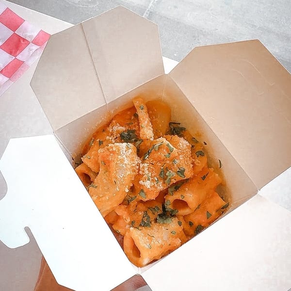 A hand holds an open white takeout box filled with rigatoni pasta in a creamy orange sauce, topped with cheese and herbs.