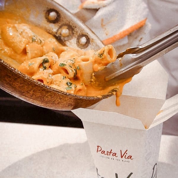 Chef using tongs to transfer creamy pasta from a pan into a "Pasta Via" takeout container.